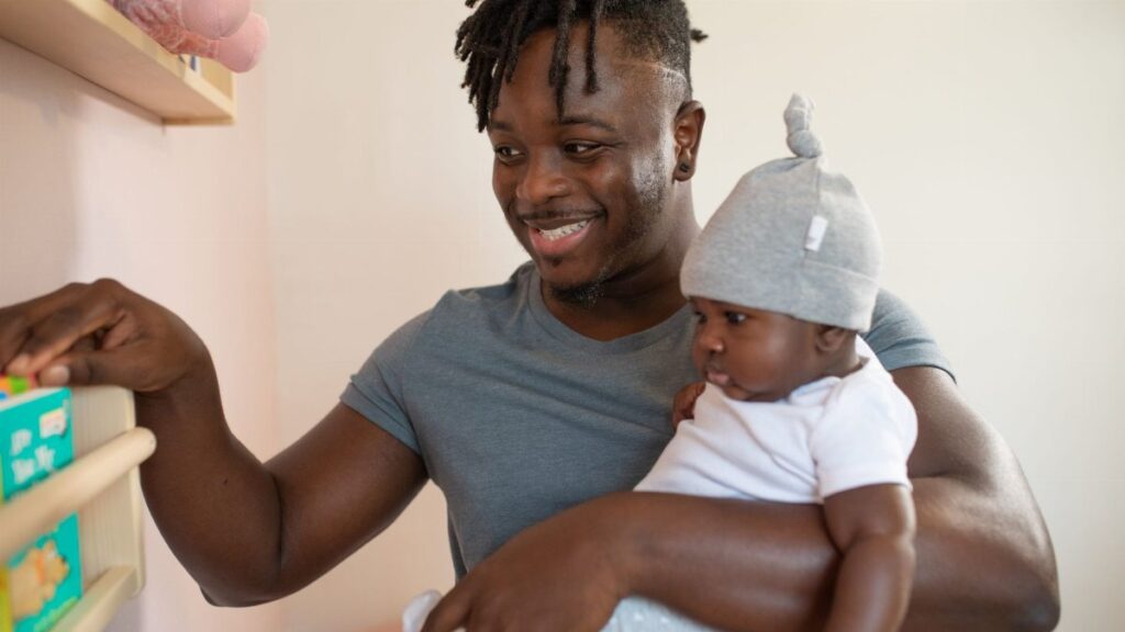 A joyful father with dreadlocks holding his baby indoors, selecting a book.