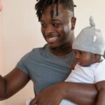 A joyful father with dreadlocks holding his baby indoors, selecting a book.