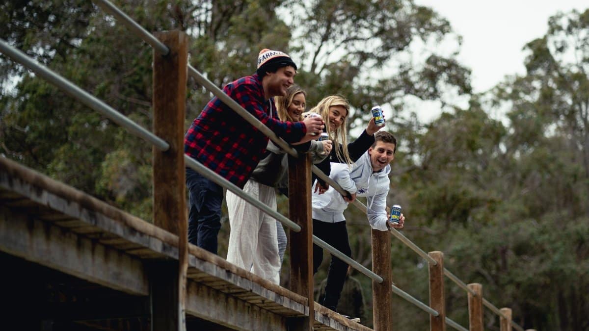 Group of friends laughing and holding beer cans on a wooden bridge in nature.