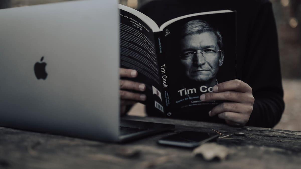 Unrecognizable ethnic male in warm clothes with opened laptop reading book and sitting outside at wooden table in autumn day