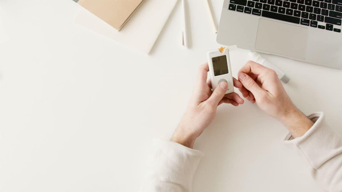 Person checking blood sugar with a glucometer on a white desk near a laptop.