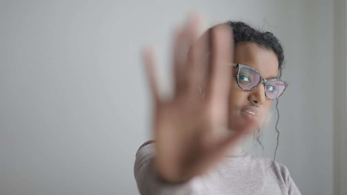 Young African American female wearing casual clothes and eyeglasses outstretching arm at camera while standing near grey wall