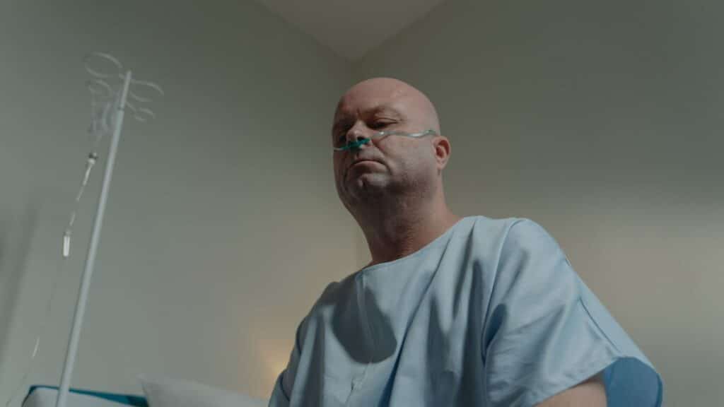 A bald man in a hospital gown sits in a room with a nasal cannula, conveying a sense of illness and solitude.