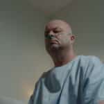 A bald man in a hospital gown sits in a room with a nasal cannula, conveying a sense of illness and solitude.