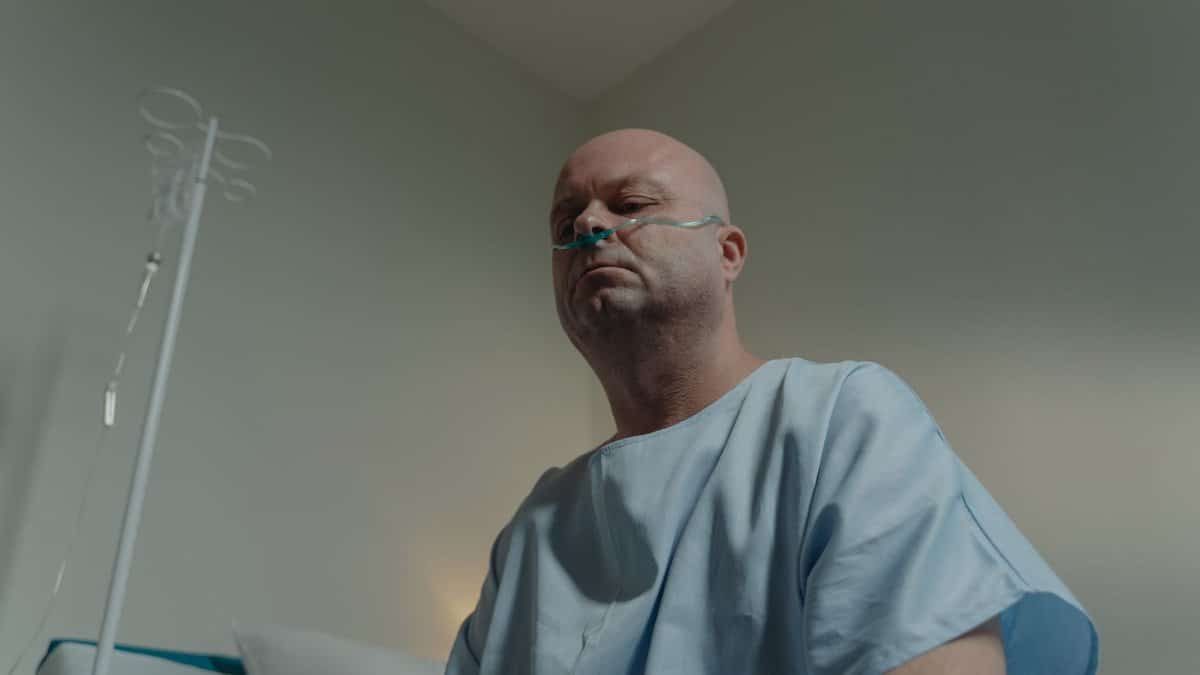 A bald man in a hospital gown sits in a room with a nasal cannula, conveying a sense of illness and solitude.