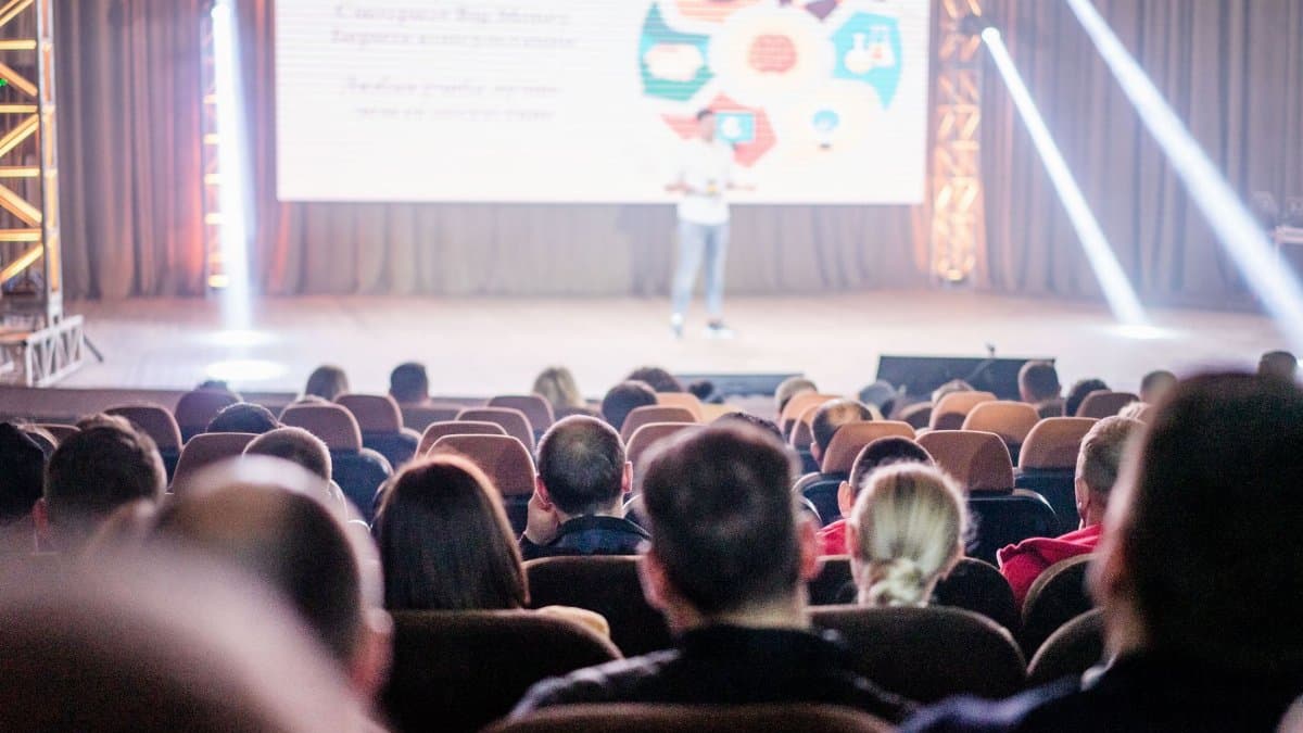 Audience attentively watching a presentation in a conference setting, with stage lights and speaker visible.