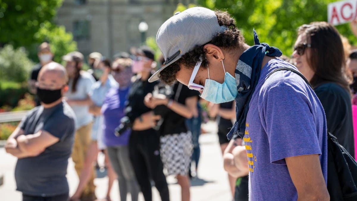 Side view of guy in casual clothes and cap and protective mask standing against crowd at sunny day