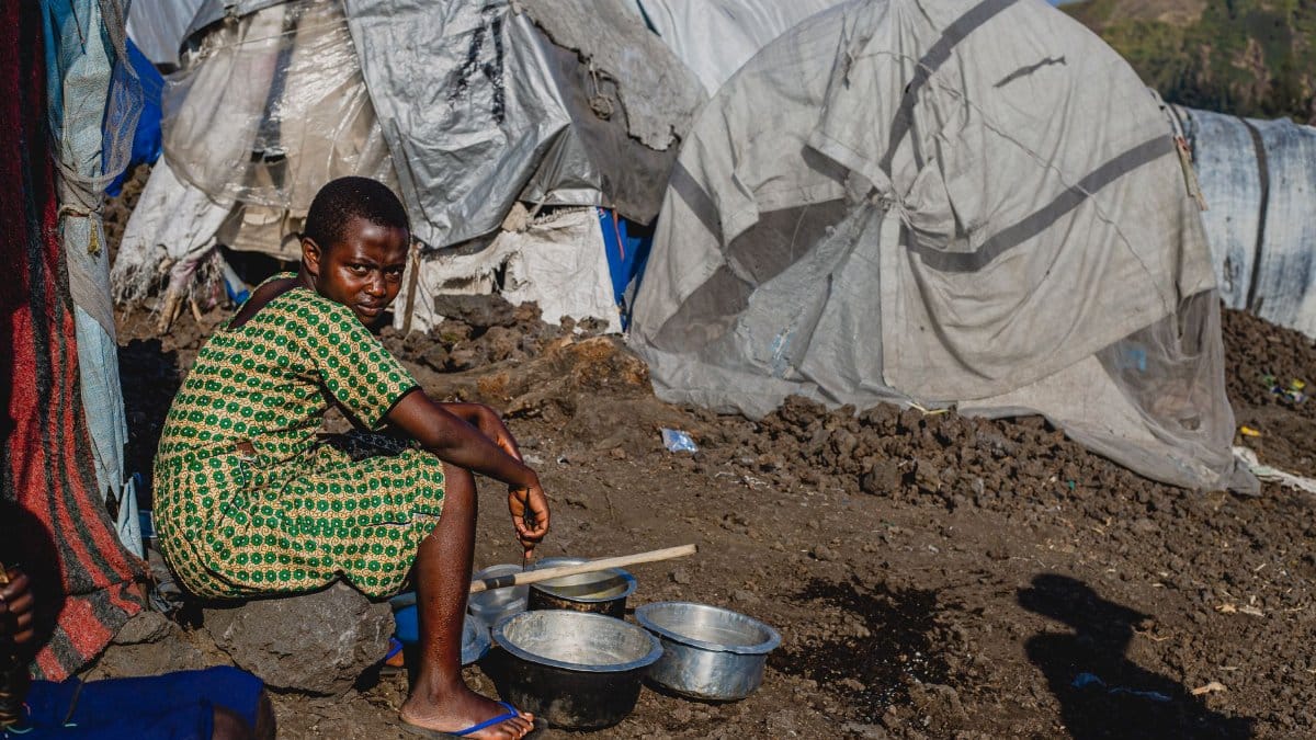 A young girl sits outside a makeshift shelter with metal bowls, highlighting resilience.