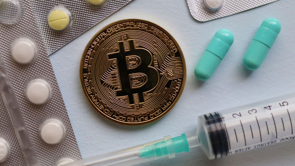 A close-up of a bitcoin, pills, and a syringe, highlighting intersections of finance and healthcare.