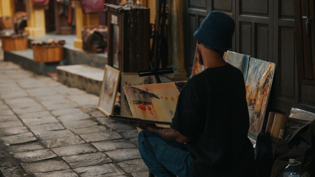 A street artist sits painting on a cobblestone street surrounded by vibrant artworks.