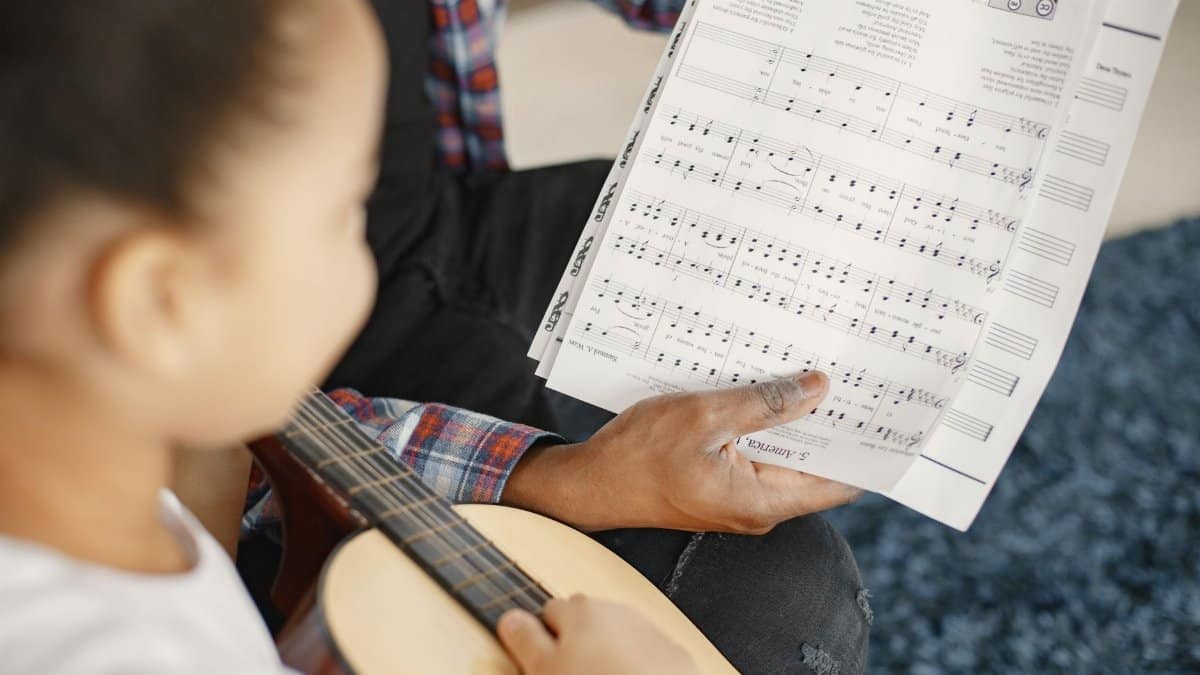A young girl learning guitar with a teacher, focusing on a music sheet.
