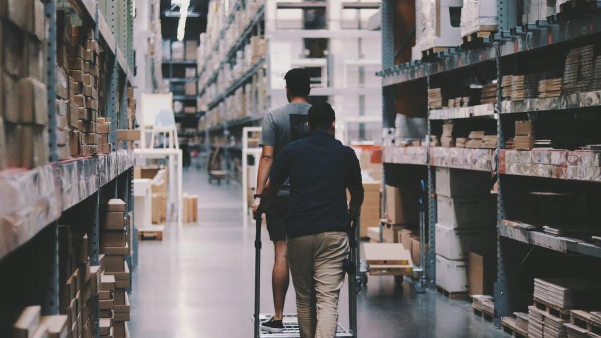 Two men maneuver a trolley in a large warehouse filled with boxes and shelves.