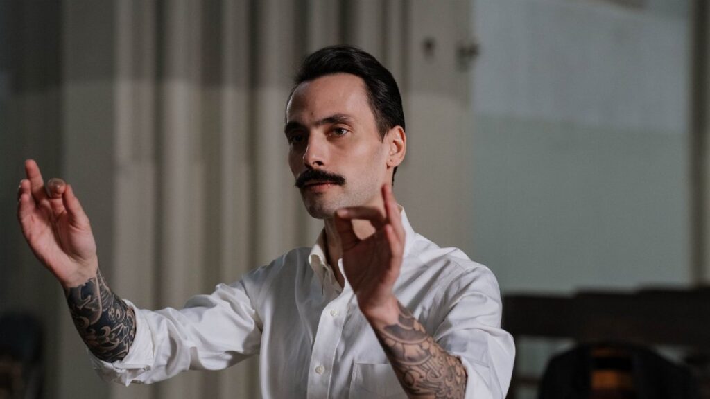 Tattooed conductor passionately leading a musical performance indoors.