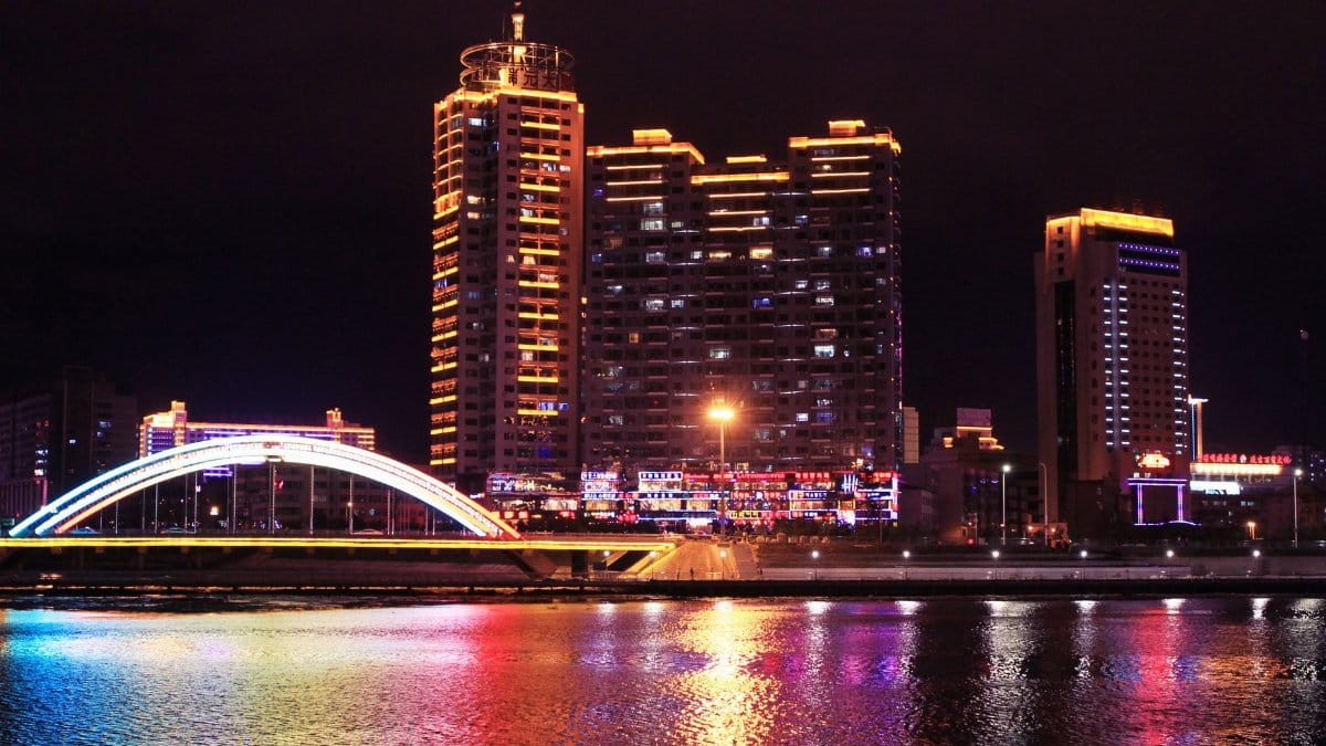 A lively cityscape featuring a bright, illuminated bridge reflected in a river at night.