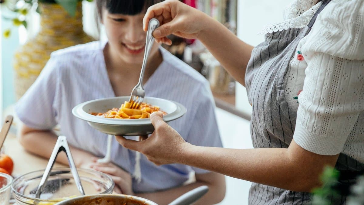 Young positive Asian females in aprons with cooked pasta with sauce in kitchen