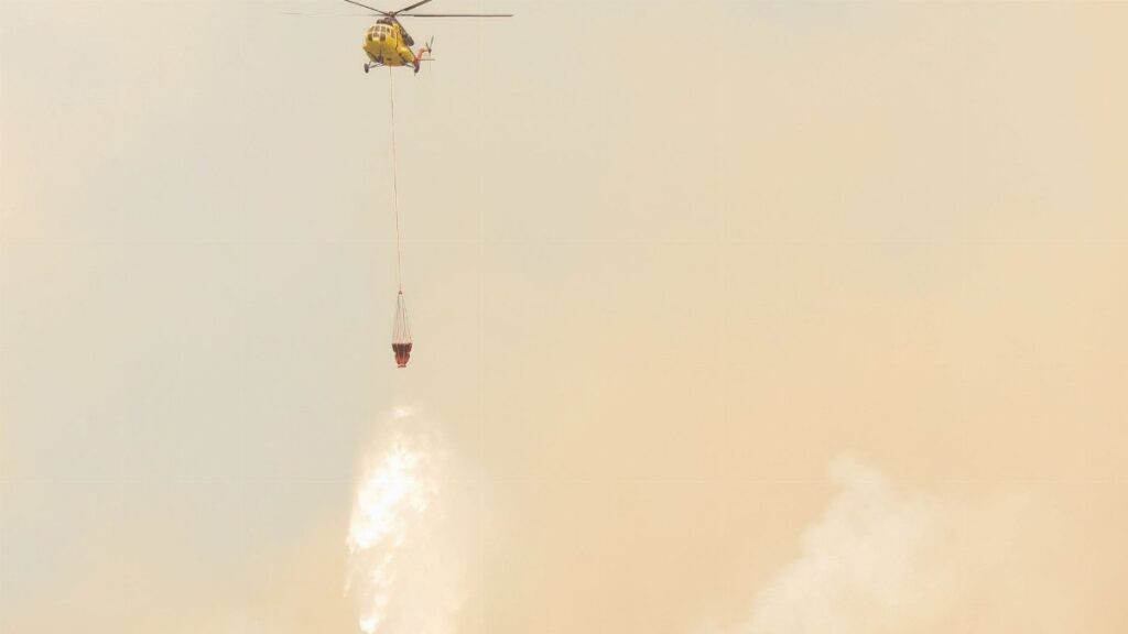 Yellow helicopter drops water to combat wildfires amidst smoky skies.