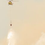 Yellow helicopter drops water to combat wildfires amidst smoky skies.