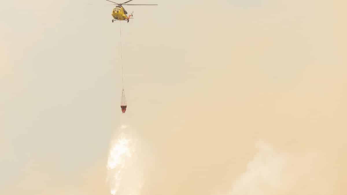 Yellow helicopter drops water to combat wildfires amidst smoky skies.