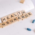 Close-up of health insurance spelled out with Scrabble tiles on a planner, alongside medication pills.