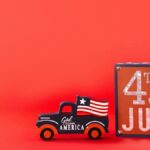 Vintage truck with American flag and 4th of July sign on red background symbolizes Independence Day celebration.