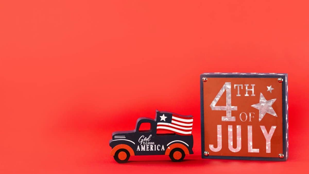 Vintage truck with American flag and 4th of July sign on red background symbolizes Independence Day celebration.