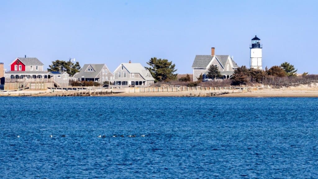 Picturesque Cape Cod lighthouse with traditional homes by the shoreline under a clear sky.