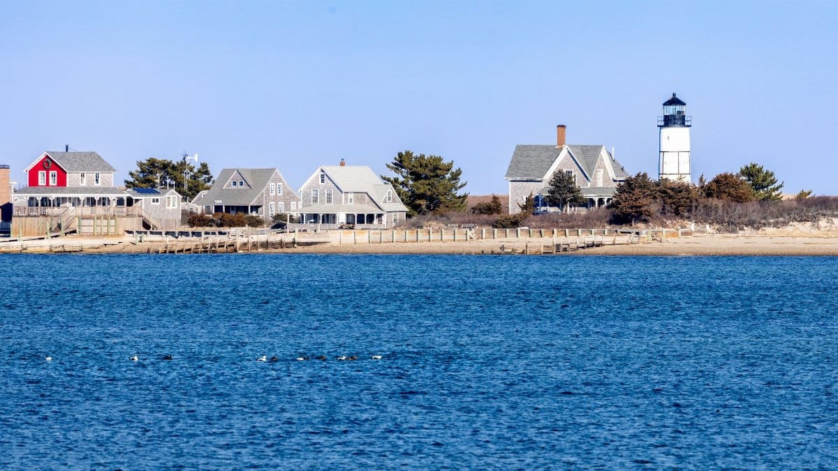 Picturesque Cape Cod lighthouse with traditional homes by the shoreline under a clear sky.