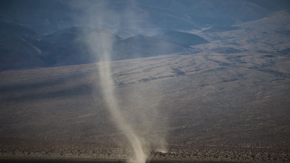 Aerial view of a dramatic dust devil swirling in a vast arid desert landscape under clear skies.