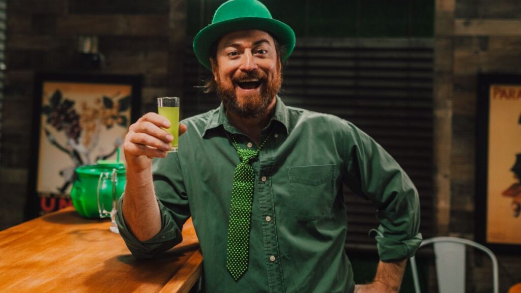Cheerful man in green attire holding a drink, celebrating St. Patrick's Day indoors.