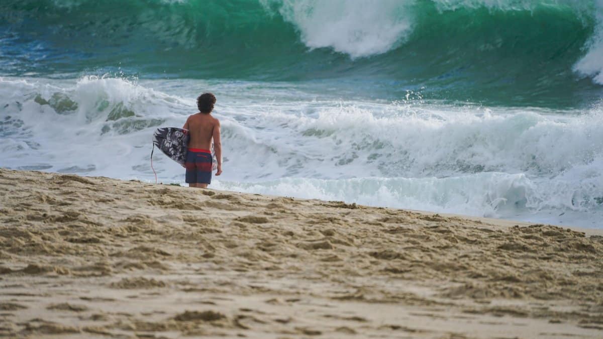 Surfer stands on sandy beach with surfboard, looking at crashing waves. Ideal for beach, surfing, and vacation themes.