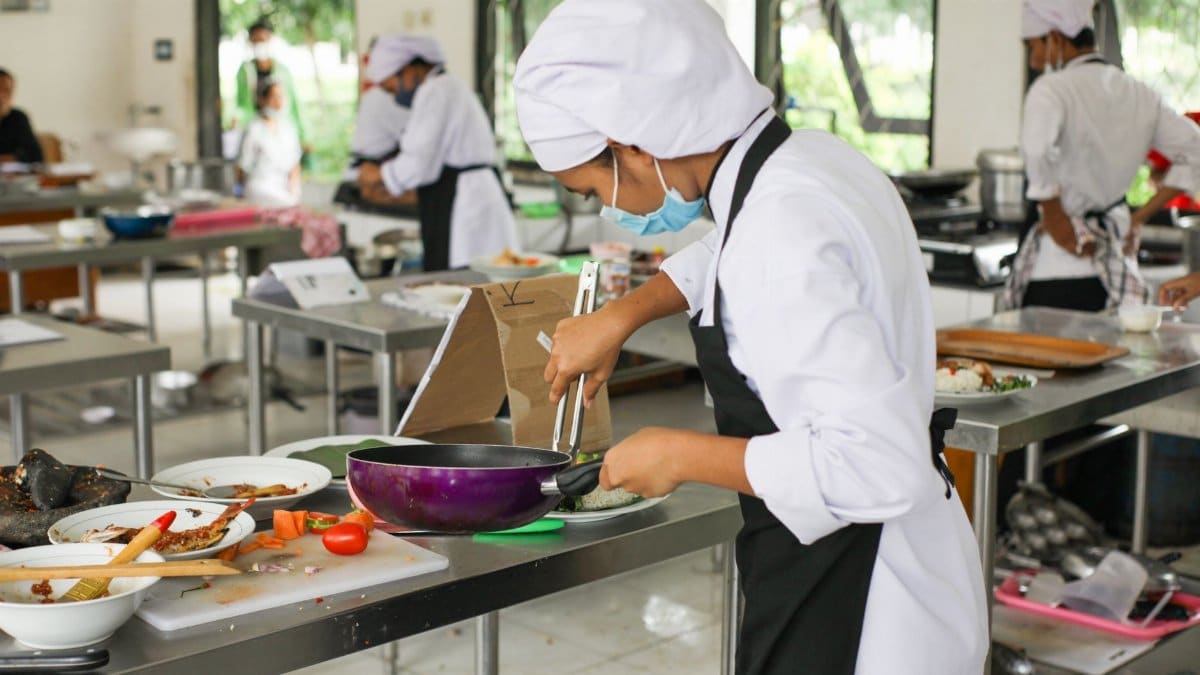 Culinary students in uniforms practice cooking skills in a professional kitchen setting.