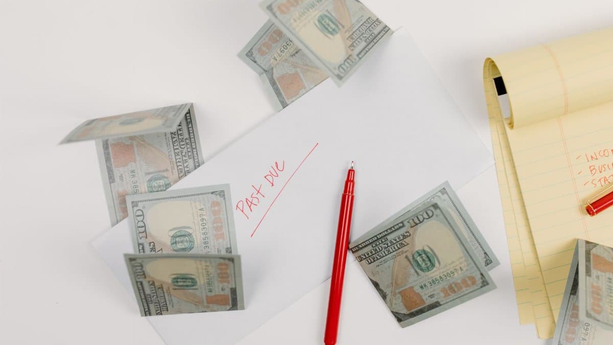 A top-down view of scattered US dollar bills with a 'past due' envelope, red pen, and notepad.