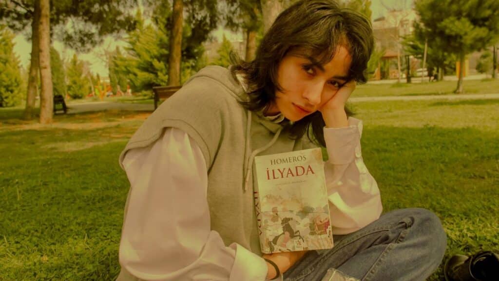 Young woman in a park reading 'Iliad' by Homer, enjoying a peaceful outdoor setting.