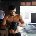 Shirtless man preparing for an intense workout session in a gym, wearing boxing gloves.