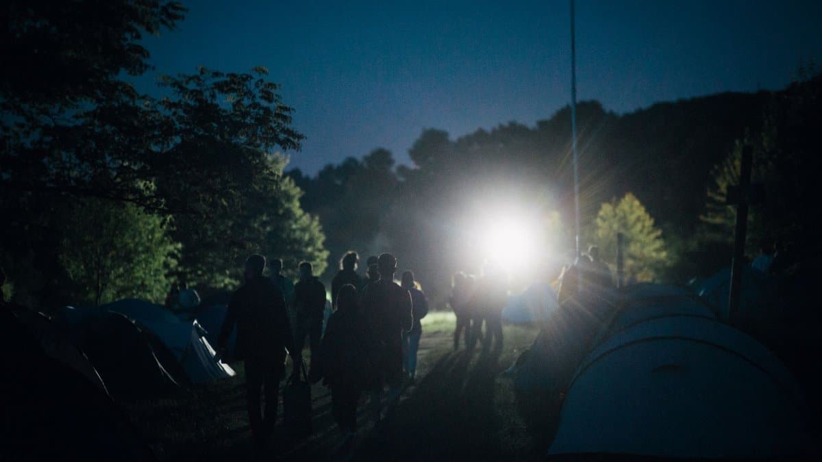 A group of people enjoying a serene camping night illuminated by distant lighting, surrounded by tents and nature.