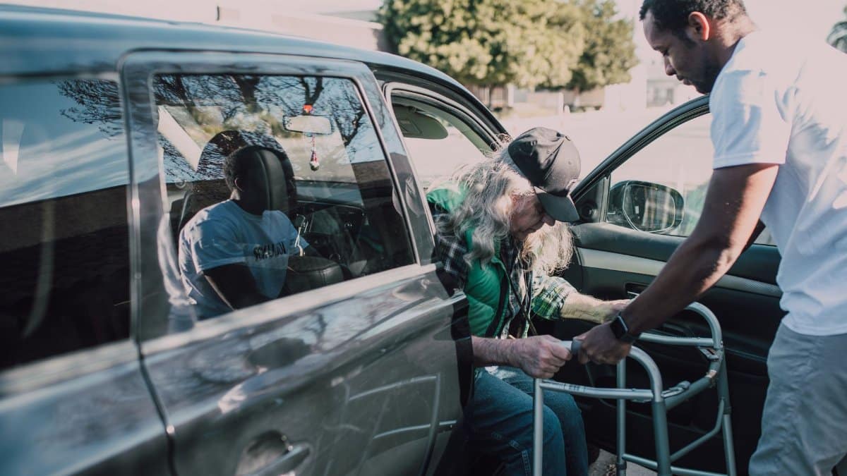Two men help an elderly person with a walker get out of a car outdoors.