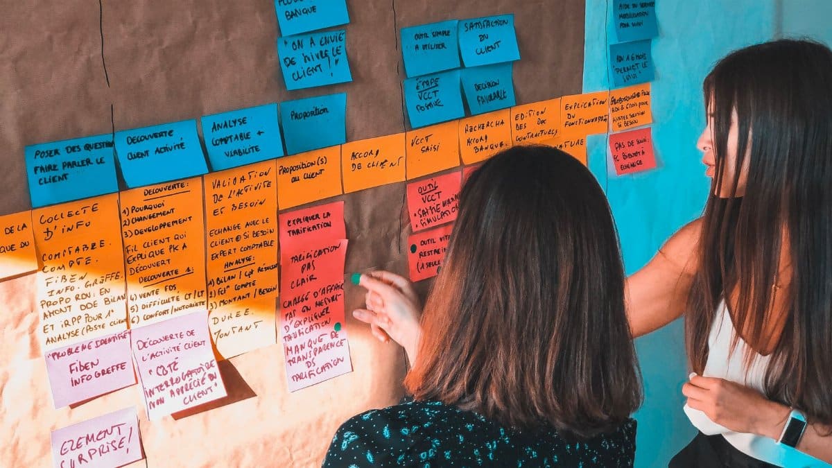 Two women engaged in collaborative planning using colorful Post-it notes on a board in a modern office space.