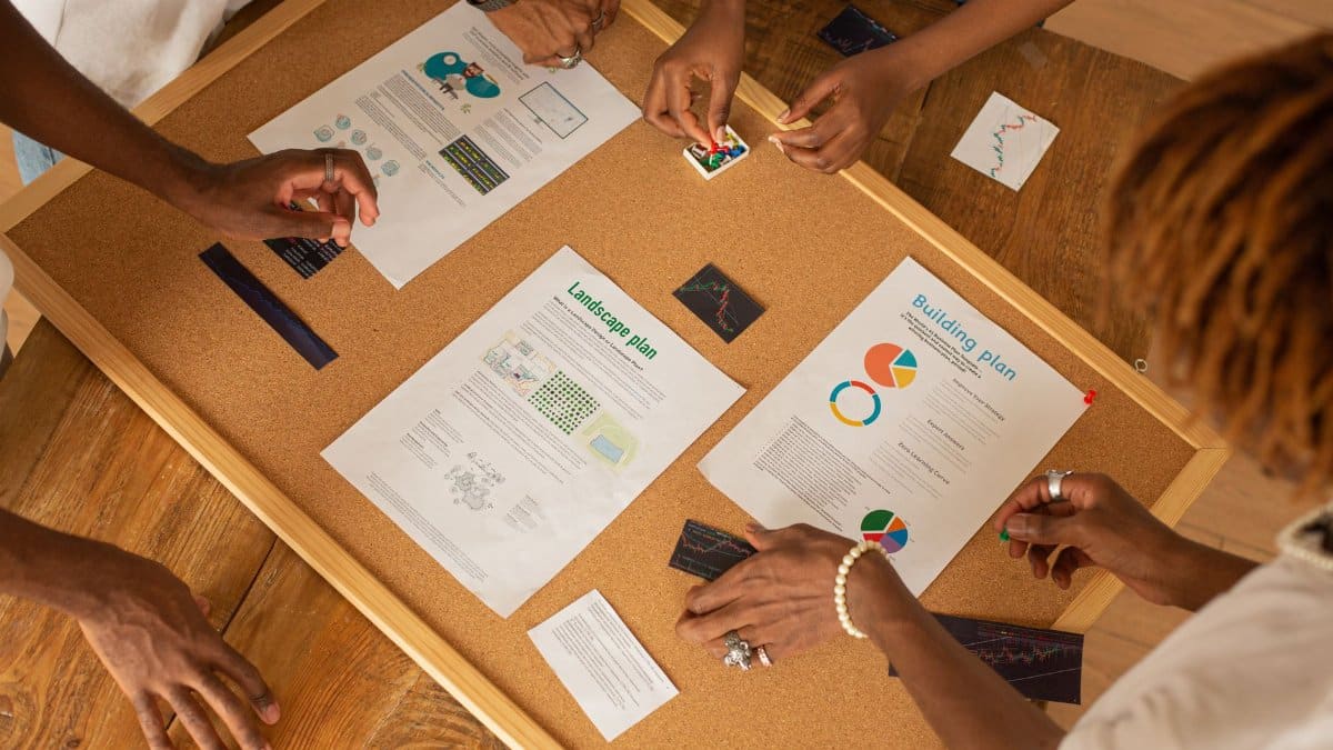 A diverse team collaborates on a workspace board with charts and plans.