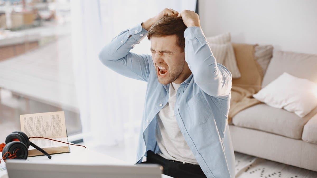 An upset man with mouth open, pulling hair at a desk with a laptop in a home setting.