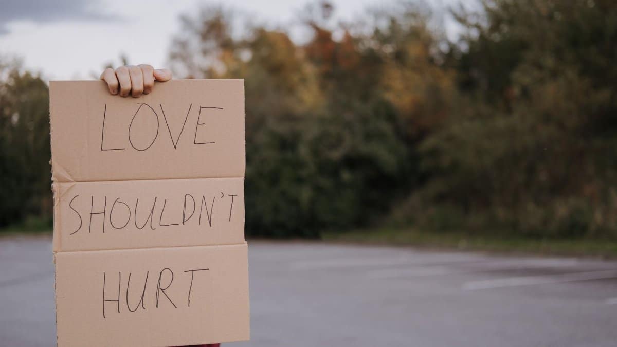 Crop anonymous person demonstrating carton signboard with slogan love should not hurt on blurred background of asphalt road and trees
