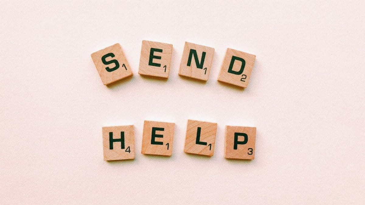 Wooden Scrabble tiles arranged to form the phrase 'send help' on a light pink background.