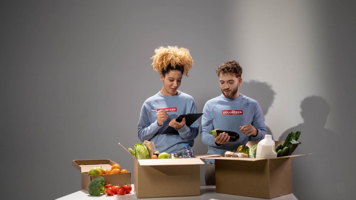 Two volunteers packing food items in cardboard boxes for community service indoors.