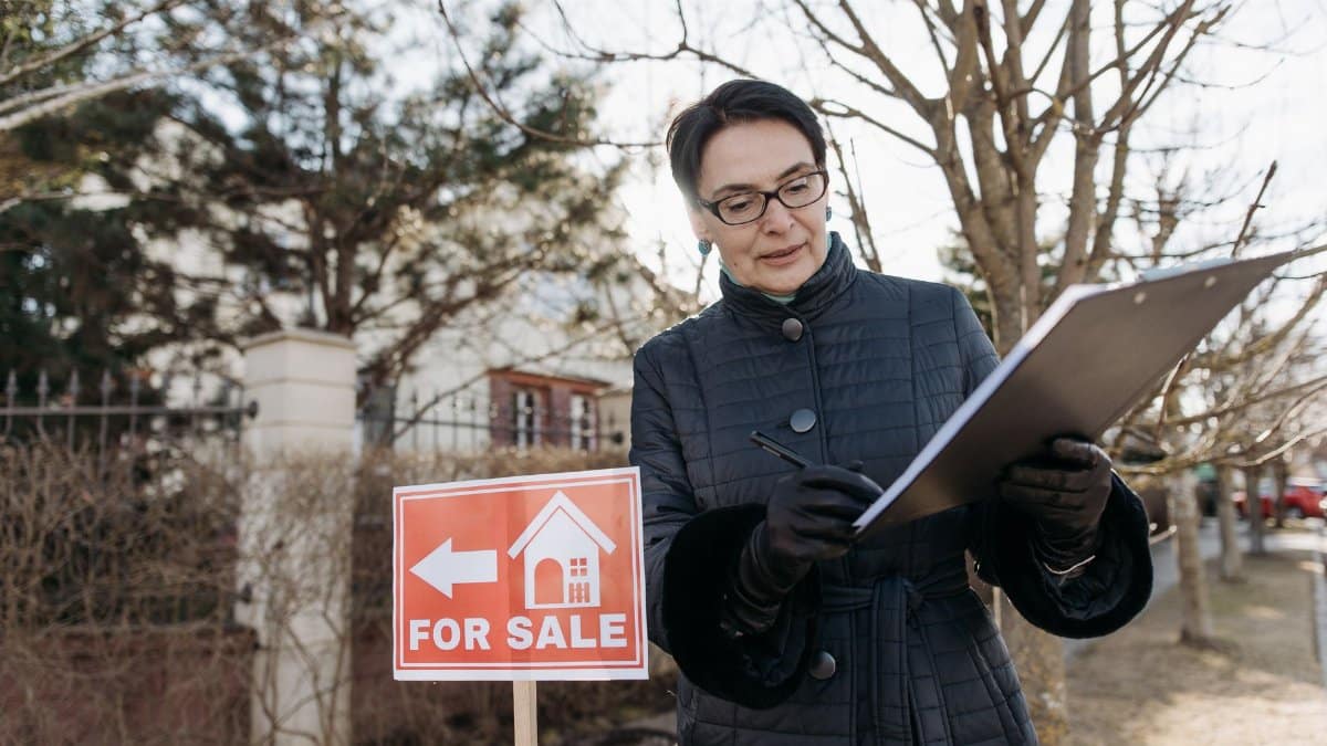 A real estate agent reviews a clipboard next to a 'For Sale' sign outside a property on a clear day.