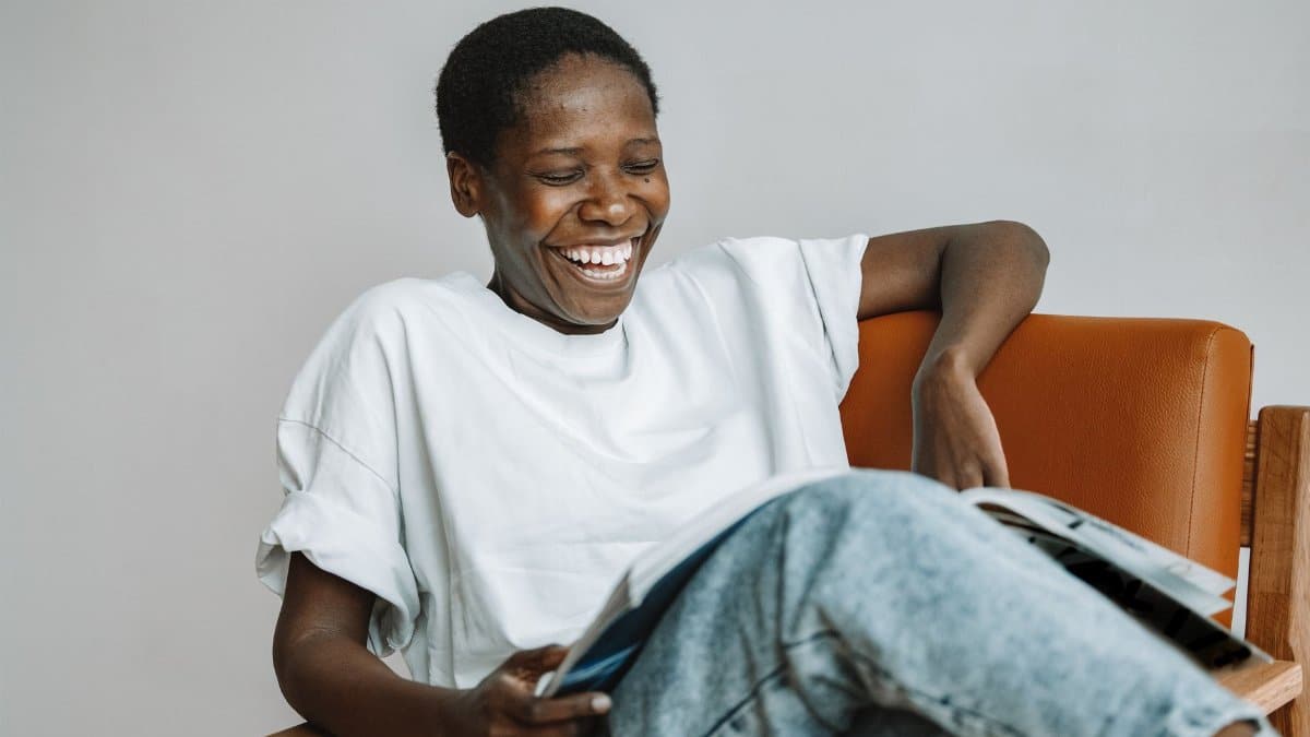 A joyful African woman laughs while reading a magazine in a cozy chair indoors.
