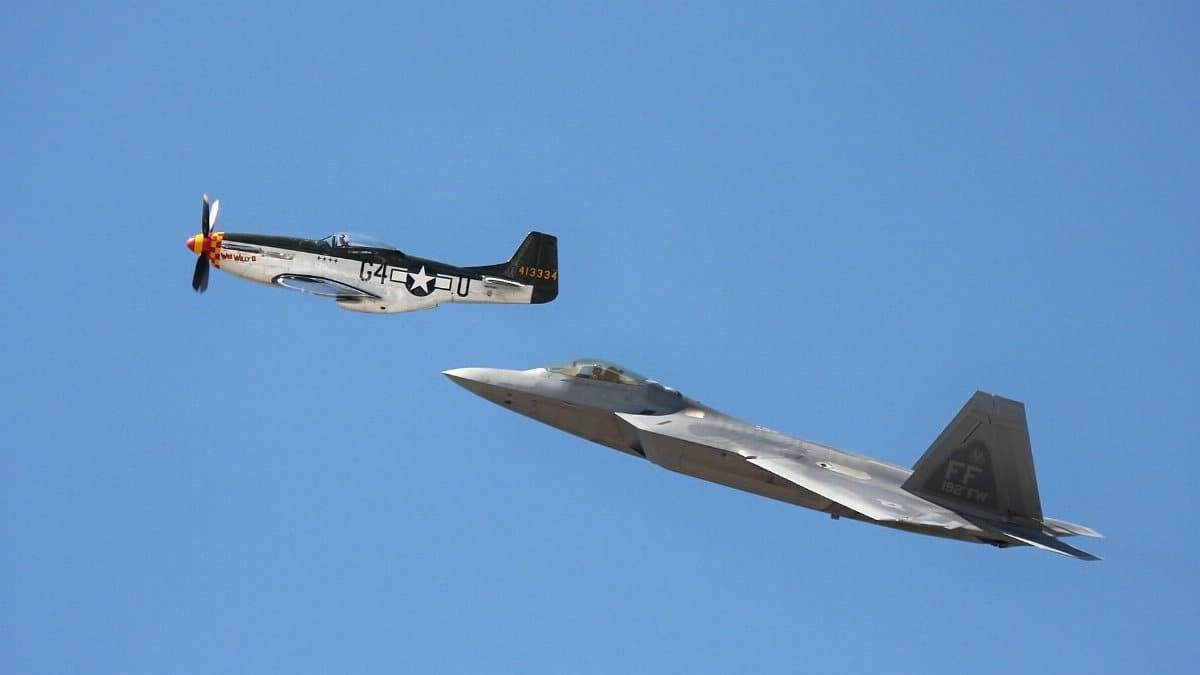 Vintage P-51 Mustang and modern F-22 Raptor in flight against clear blue sky.