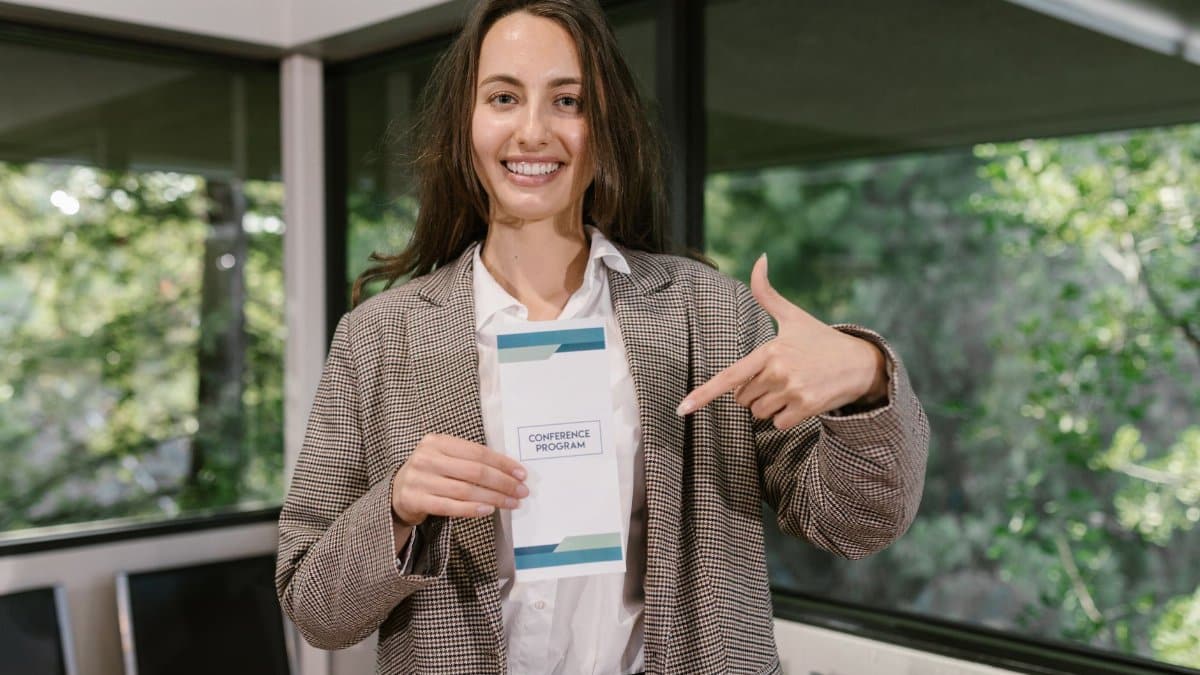 Woman indoors holding and pointing at a conference program pamphlet with a smile.