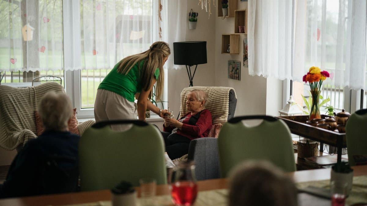 Caregiver assisting elderly women with tea in a cozy nursing home setting.