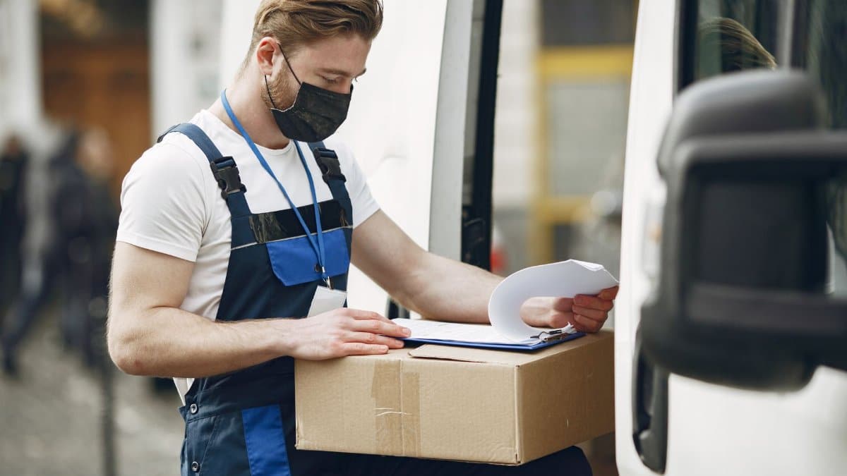A delivery worker wearing a face mask checks a package next to a van, ensuring safety measures.