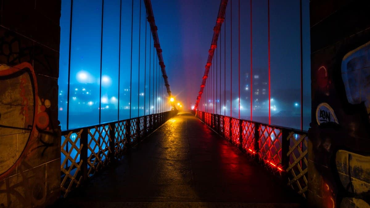 A neon-lit suspension bridge glowing in misty conditions during the night in Glasgow.