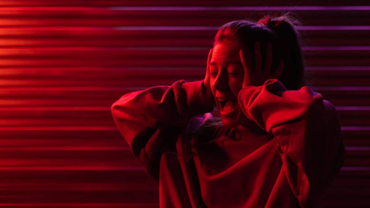 Crying frustrated woman with eyes closed in hoodie in bright ultraviolet light in darkness
